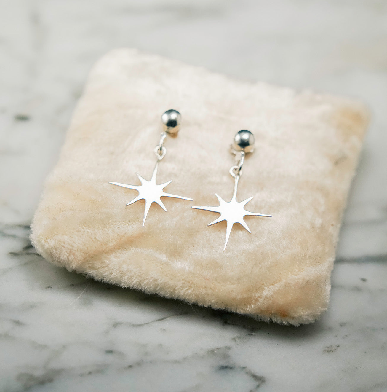 
                  
                    Silver star-shaped earrings on a beige fabric surface with a marble background
                  
                